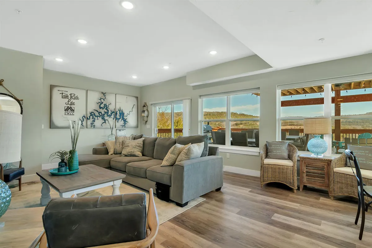 This interior image showcases a welcoming great room featuring a large sectional sofa, decorative wall art, woven chairs with cushions, a coffee table adorned with decorative items, and expansive windows overlooking the picturesque landscape. The room exudes coziness and is perfect for social gatherings.