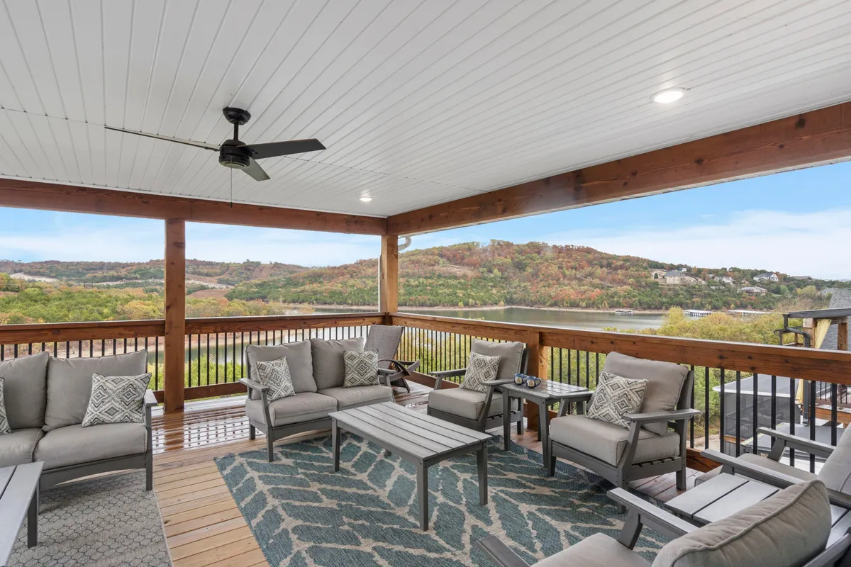 The image showcases the exterior of a spacious, beautifully furnished lakefront home with a comfortable seating area under exposed wooden beams, a decorative rug, and sweeping panoramic views of Table Rock Lake. The cozy lounge space is perfect for relaxing or entertaining, with spectacular sunset vistas in the background, evoking a serene and inviting mood.