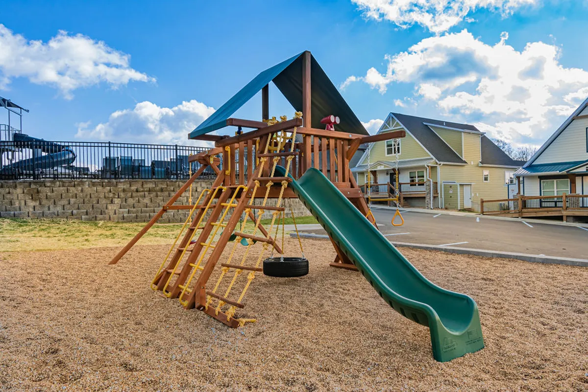 A cheerful playground area featuring a modern slide and swing set, with children playing and families interacting. The surrounding green space suggests a safe, inviting area for kids to explore and parents to connect.