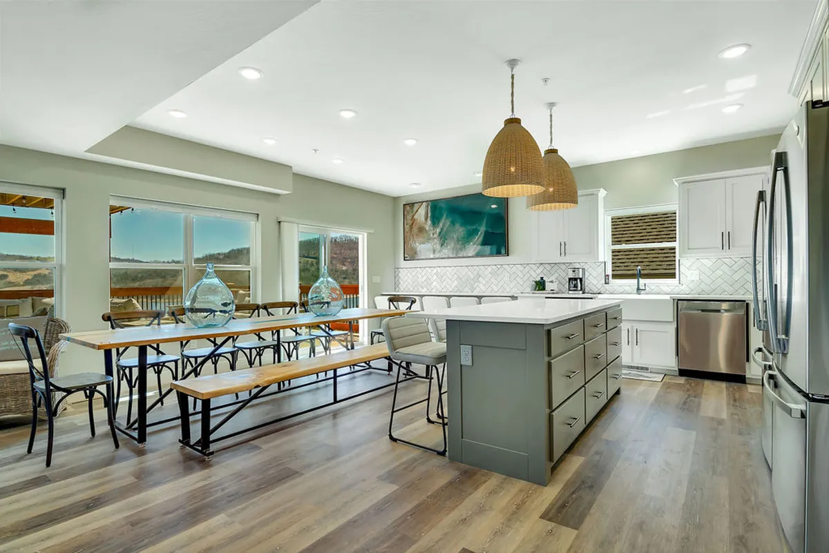 This image showcases a spacious kitchen with a large central island, stylish pendant lighting, an inviting dining table, and expansive windows that flood the space with natural light. The kitchen appears well-equipped for group meal preparation and entertaining, perfectly illustrating a modern chef's domain.