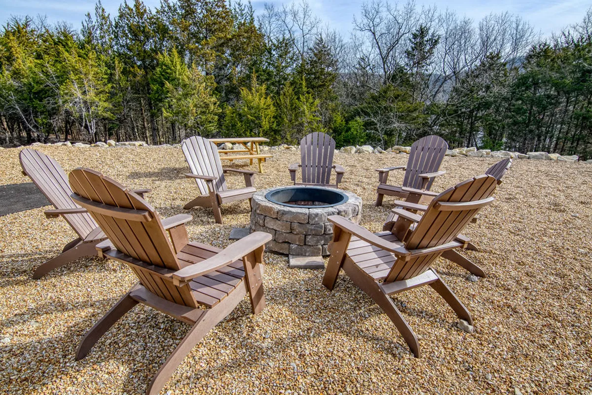 A cozy fire pit surrounded by Adirondack chairs nestled among trees, inviting guests to gather in the evening. The setup suggests storytelling, making s’mores, and stargazing under the night sky.
