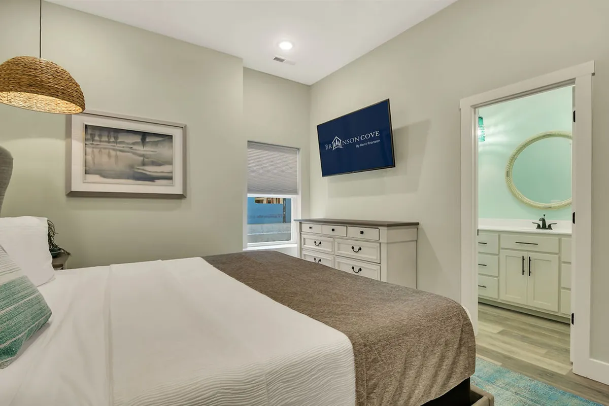 A serene bedroom featuring a bed with white linens and a brown throw blanket, a woven pendant light overhead, a television mounted on the wall, a dresser with multiple drawers, and a doorway leading to a private bathroom. Soft natural light fills the room, creating an inviting atmosphere ideal for relaxation.