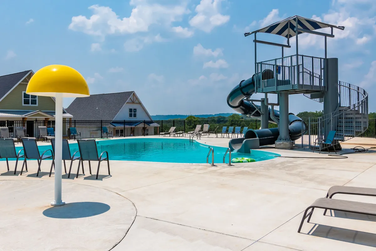 This image features a lively outdoor swimming pool with a prominent water slide, yellow umbrella, lounge chairs, and a clubhouse in the background. Families and friends are enjoying themselves by the poolside, suggesting both relaxation and active fun.