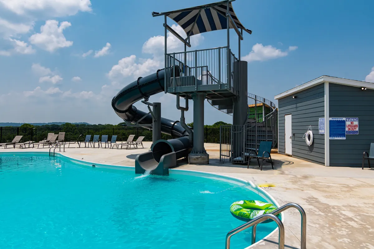 An outdoor swimming pool complete with a water slide, lounge chairs, and a lifeguard station is pictured. The pool area is lively and basked in sunlight, suggesting family-friendly, summer fun.