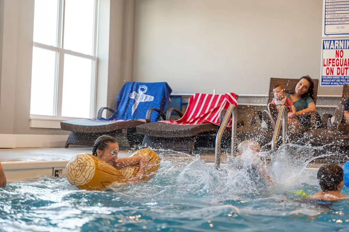 Children joyfully play in a bright indoor swimming pool, surrounded by loungers and a colorful floatation device. Large accessible poolside space is visible, providing a safe area for swimmers and those simply relaxing.