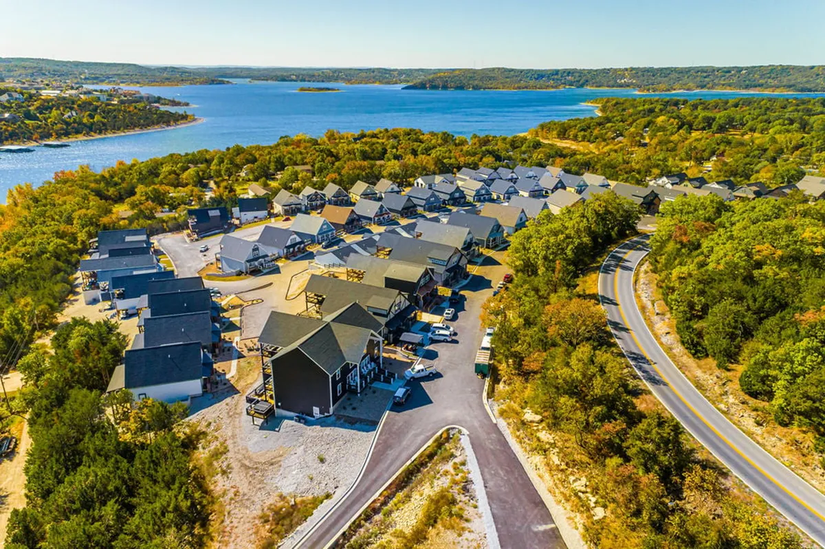 A serene aerial view overlooking Table Rock Lake, with the water, a residential area, a tree line, and a road visible. The scene conveys community, natural beauty, and opportunities for boating, hiking, and gathering by the water. The landscape suggests a space for both active and relaxing outdoor pursuits.