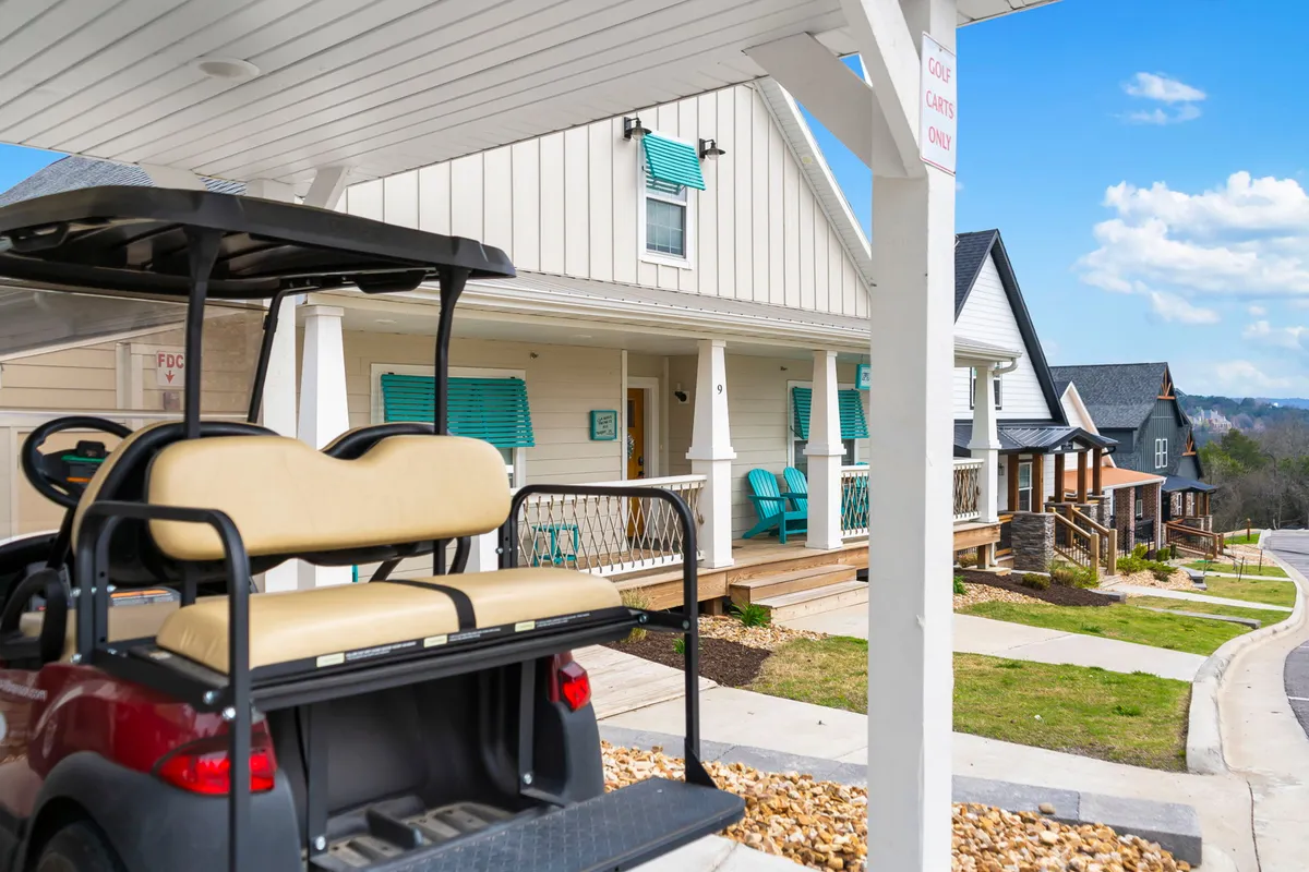 The image features a cheerful exterior scene with a golf cart parked near colorful homes with inviting front porches and shutters. The setting suggests easy access to golf and a relaxed community feel, with porches perfect for morning coffee or unwinding after a day exploring Branson.