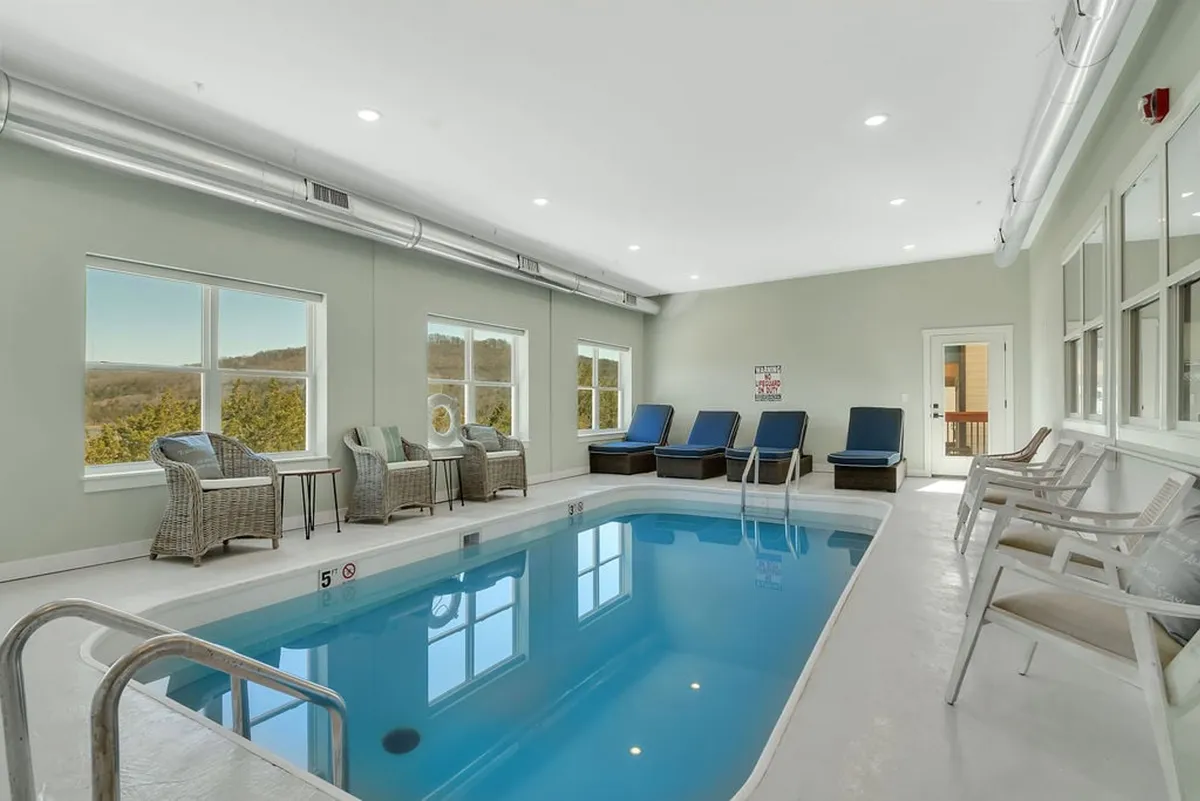 A bright, tranquil indoor swimming pool surrounded by large windows that fill the space with natural light. Comfortable lounge chairs are arranged poolside, providing a relaxing area to supervise children or unwind after a swim. The inviting atmosphere is perfect for both active play and peaceful relaxation.
