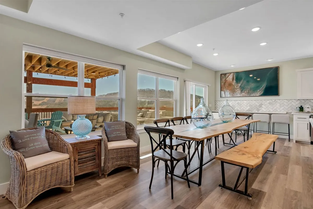 A spacious dining area anchors this scene with a large dining table surrounded by wicker chairs, decorative vases, and a beautiful view framed by the windows. The setting is perfect for shared meals, conversations, or group celebrations.