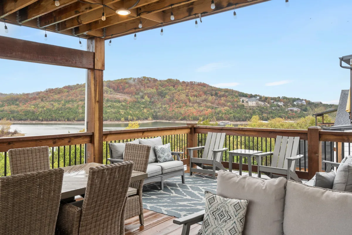 A welcoming outdoor space with a dining table set up for social gatherings, comfortable chairs, and a sweeping scenic view of hills and water. String lights add to the inviting evening ambiance, perfect for alfresco meals.